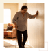 An elderly woman leans against a wall for support while walking with a cane in a living room.