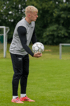 Ein blonder junger Mann in Sportkleidung hält einen Fußball auf einem grünen Feld.