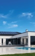 Modernes weißes Haus mit Solarmodulen auf dem Dach, Terrasse, Pool und Liegestühlen unter blauem Himmel.