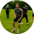 Fußballspieler in schwarzem Trainingsanzug beim Üben auf dem Rasenplatz, weitere Personen im Hintergrund.