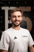 Lächelnder Physiotherapeut im weißen Adidas-Shirt mit dem Logo „THERAPIER this is PHYSIO“ vor dunklem Hintergrund und Pier5.