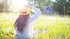 Frau mit Strohhut und weißer Bluse sitzt von hinten gesehen in einer sonnigen Blumenwiese mit Gänseblümchen.