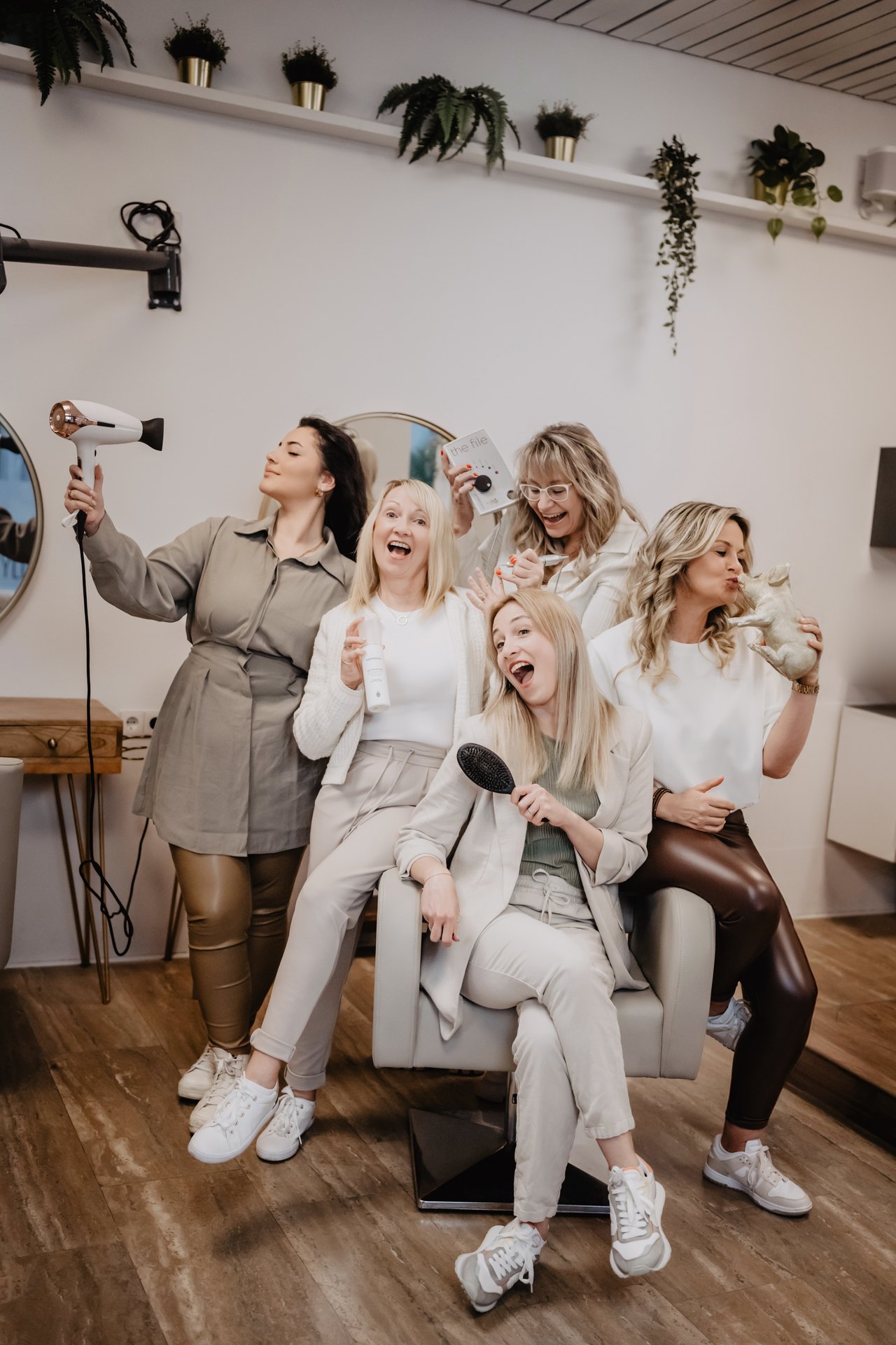 Fünf lächelnde Friseurinnen posieren mit Föhn, Haarbürste und Pflegeprodukten in einem modernen Friseursalon.