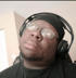 Man in black durag and t-shirt, wearing glasses and headphones, looking up and to the right.
