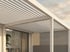 Weiße Pergola mit Lamellendach über einer Terrasse vor einem Haus mit Glasfront.
