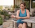 A woman in a blue tank top sits at an outdoor table and holds her forearm with a pained expression.