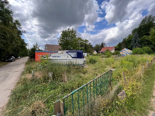 Ein Boot und ein oranger Container stehen auf einer grünen Wiese neben einem Feldweg unter bewölktem Himmel.