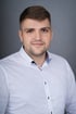 Ein Mann mit kurzem braunem Haar und Bartansatz trägt ein weißes Hemd mit gemustertem Kragen.