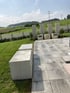 Moderne Terrasse mit grauen Steinplatten, Betonquadern als Sitzgelegenheit und hohen Sichtschutzelementen vor grüner Wiese.