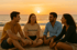 Vier Freunde sitzen am Strand und unterhalten sich bei Sonnenuntergang; Meer im Hintergrund.