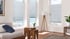 Drei Fenster mit grauen Plissees in einem Raum mit Blick auf einen Strand und das Meer.