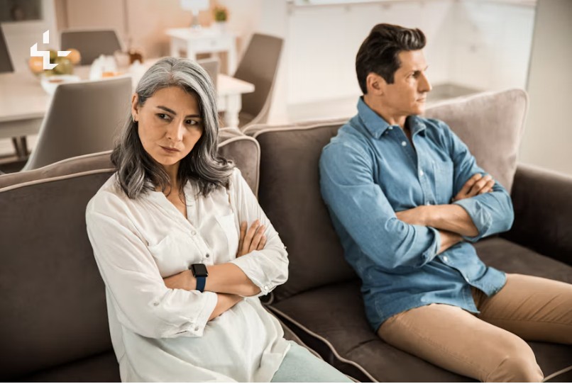 Eine Frau um die 50 sitzt mit verschränkten Armen auf dem Sofa, ihr Mann schaut in die andere Richtung
