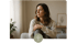 Woman in a light sweater and gray pants, sitting on a sofa, looking out a window.