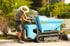 Jens Gandyra mit Strohhut bedient einen hellblauen Messersi TC85d Kettendumper bei Gartenarbeiten auf einem Gehweg.