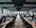Großraumbüro mit Reihen von Mitarbeitern an Computern, schwarzer Decke und großen Fenstern.