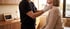 A healthcare professional in black scrubs and gloves performs a physical neck examination on a seated patient.