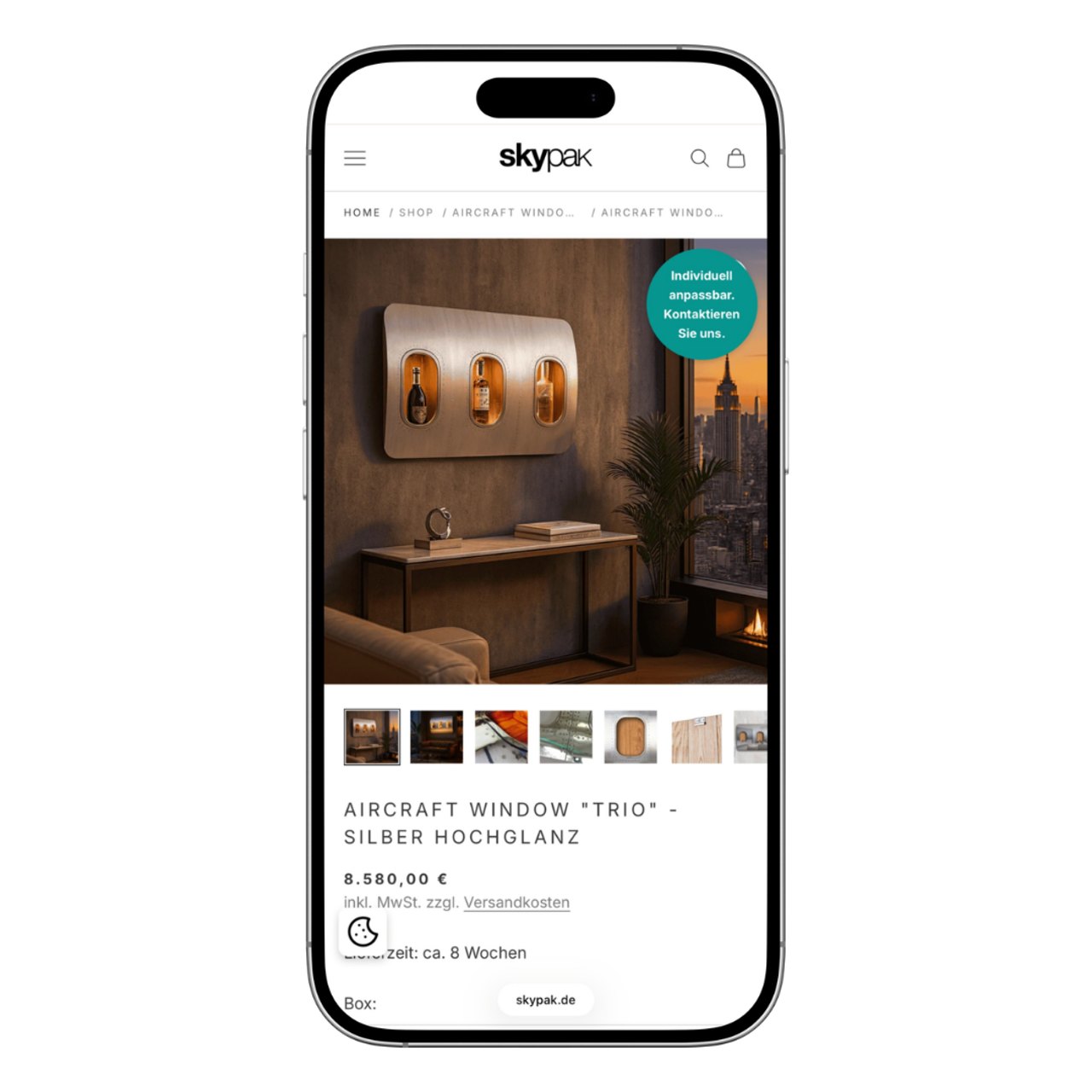 Ein Smartphone zeigt die SkyPak-Website mit einem silbernen Hochglanz-Flugzeugfenster "Trio" als Wandregal.