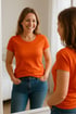 Eine lächelnde Frau in einem orangefarbenen T-Shirt und Jeans steht vor einem Spiegel.