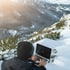 Mann mit Mütze und Jacke tippt auf Laptop in verschneiter Berglandschaft.