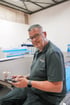 Andreas Singer mit Brille und grauem Poloshirt bedient ein Smartphone in einer Werkstatt neben einer Kaffeetasse.