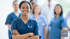 Smiling female nurse in blue scrubs with a stethoscope around her neck, arms crossed, with medical team blurred behind.