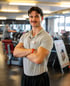 Ein lächelnder Fitnesstrainer mit verschränkten Armen steht in einem Fitnessstudio.