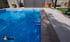 Ein klarer blauer Pool mit Mosaikfliesen neben einer grauen Terrasse und einem geschlossenen Tor im Hintergrund.