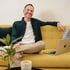 Lächelnder Mann sitzt mit Laptop und Kaffee auf gelbem Sofa und schaut in die Kamera.