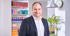 Ein lächelnder Mann in Anzug und weißem Hemd steht vor einem Bücherregal mit roten und blauen Ordnern.