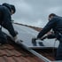 Zwei Handwerker in dunkler Arbeitskleidung montieren eine Photovoltaikanlage auf einem Ziegeldach unter bewölktem Himmel.