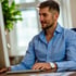 Junger Mann in blauem Hemd arbeitet konzentriert am Computer, sonniges Fenster im Hintergrund.