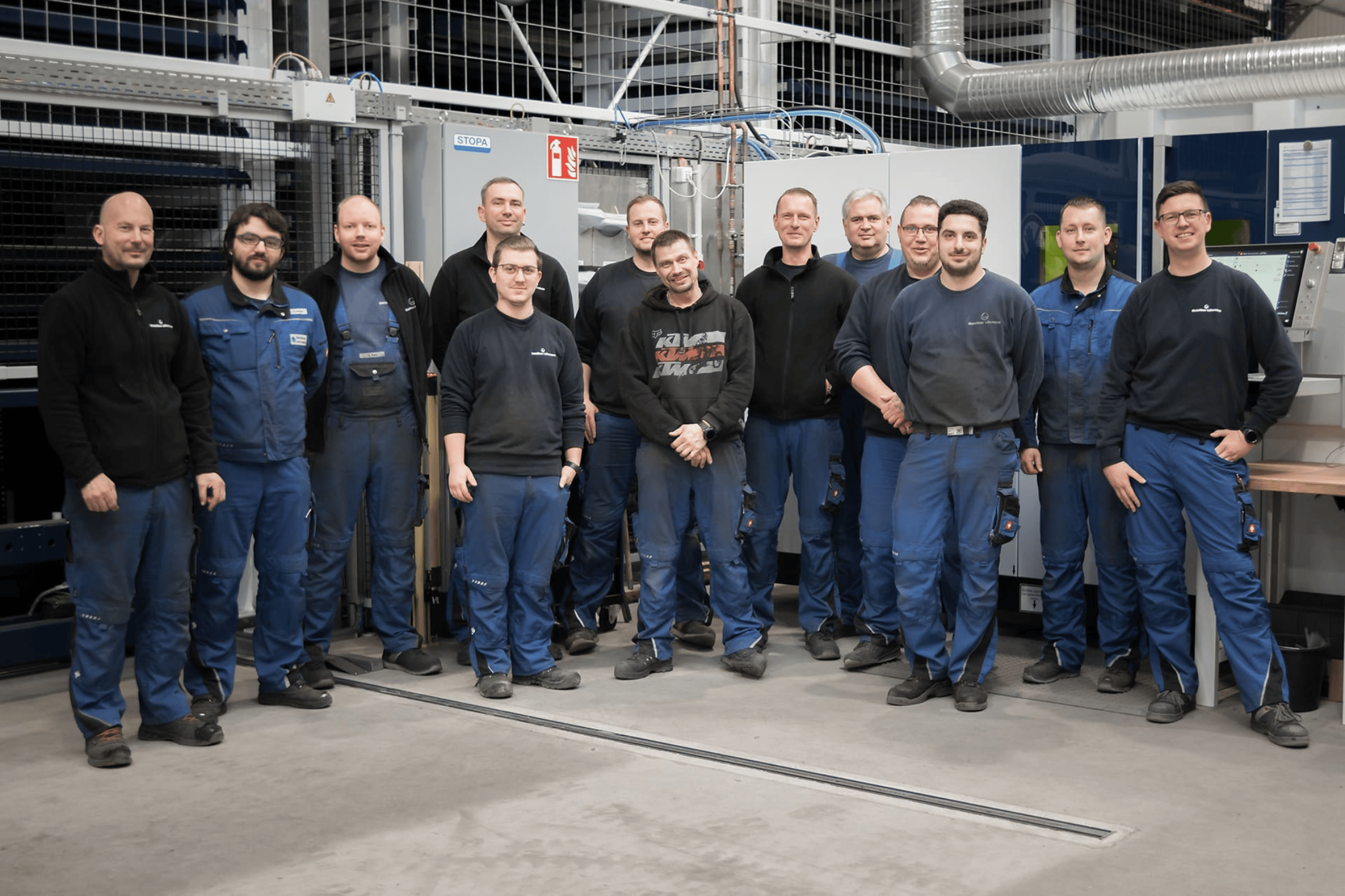 Eine Gruppe von 13 Männern in Arbeitskleidung posiert lächelnd in einer Fabrikhalle vor Maschinen.