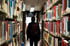 Person mit Rucksack steht von hinten betrachtet in einem schmalen Gang zwischen hohen Bücherregalen einer Bibliothek.