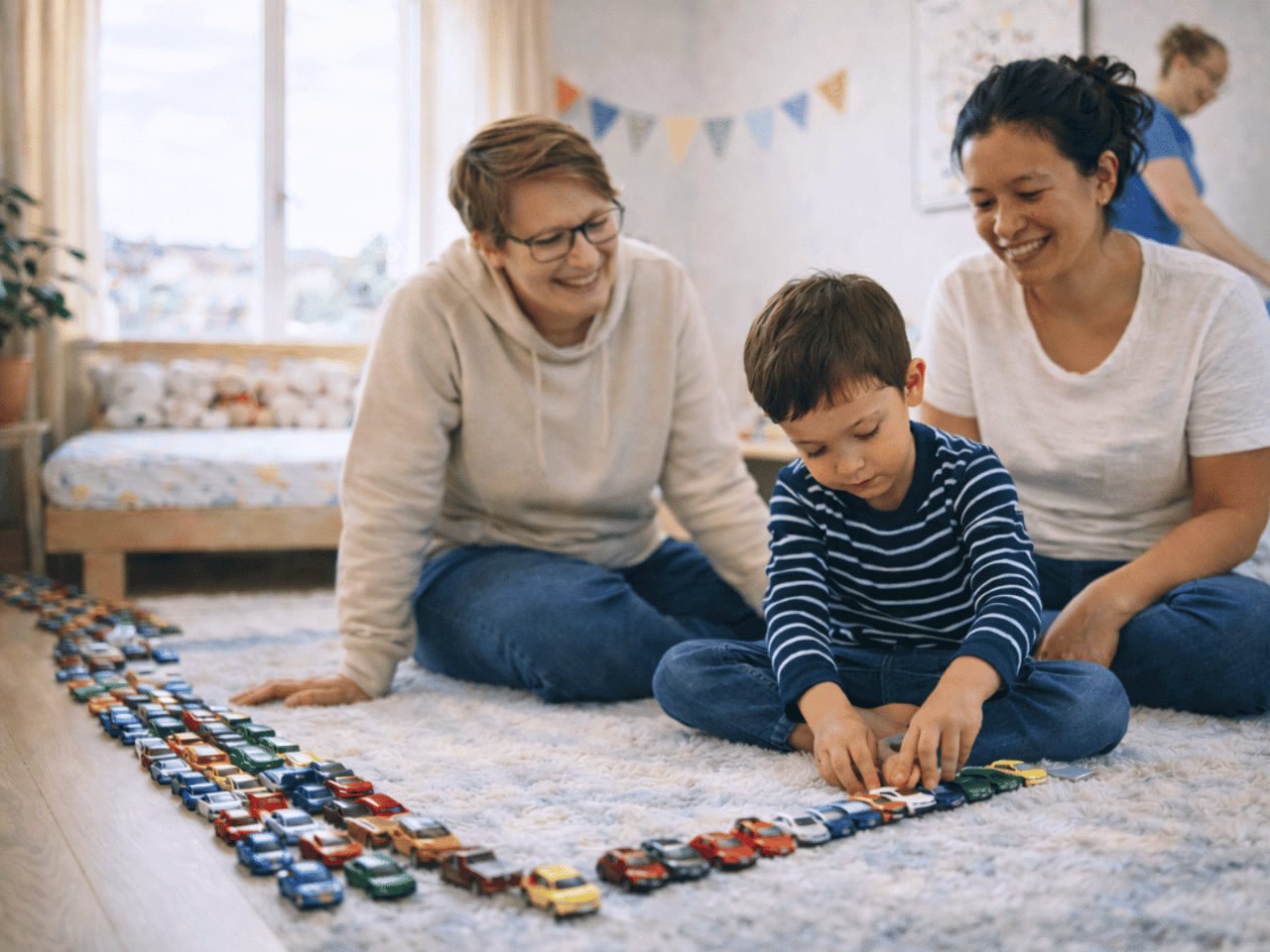 Zwei Frauen spielen mit einem Baby auf dem Boden, während eine Frau im Hintergrund ein Zimmer aufräumt.