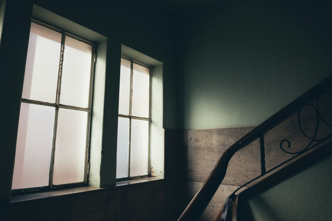 Ein stilles Altbau-Treppenhaus mit hohen Sprossenfenstern und geschwungenem Metallgeländer im gedämpften Tageslicht.