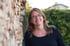 Frau mit Brille lächelt neben einem Stapel Holzscheite, im Hintergrund ein Haus und grüne Büsche.