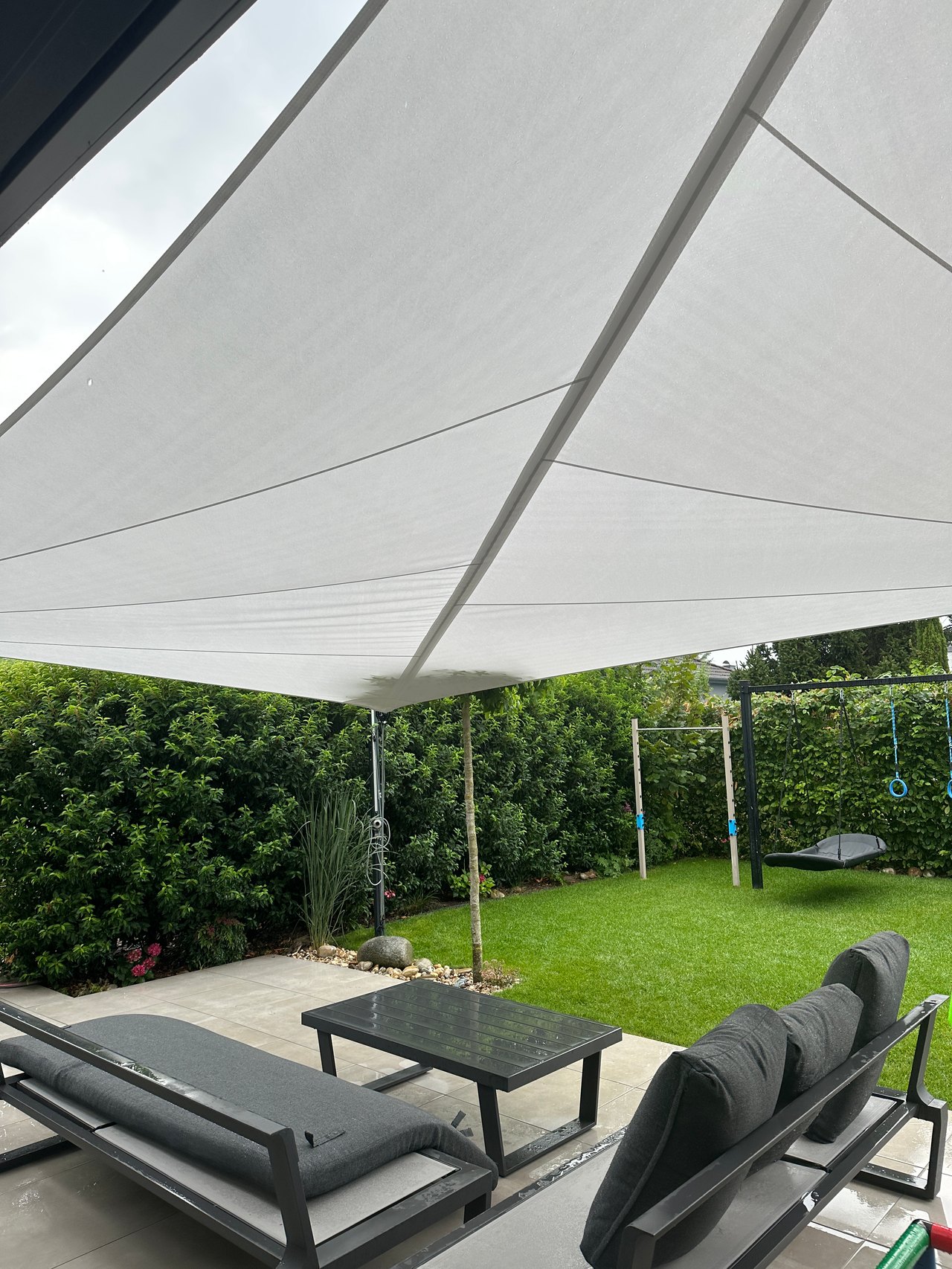 Lounge-Sitzgruppe unter einem hellen Sonnensegel vor Rasen und Hecke