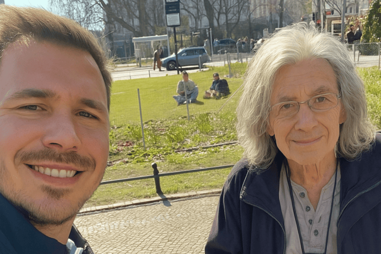 Zwei Männer lächeln in die Kamera, einer mit kurzem blondem Haar, der andere mit langen grauen Haaren und Brille.