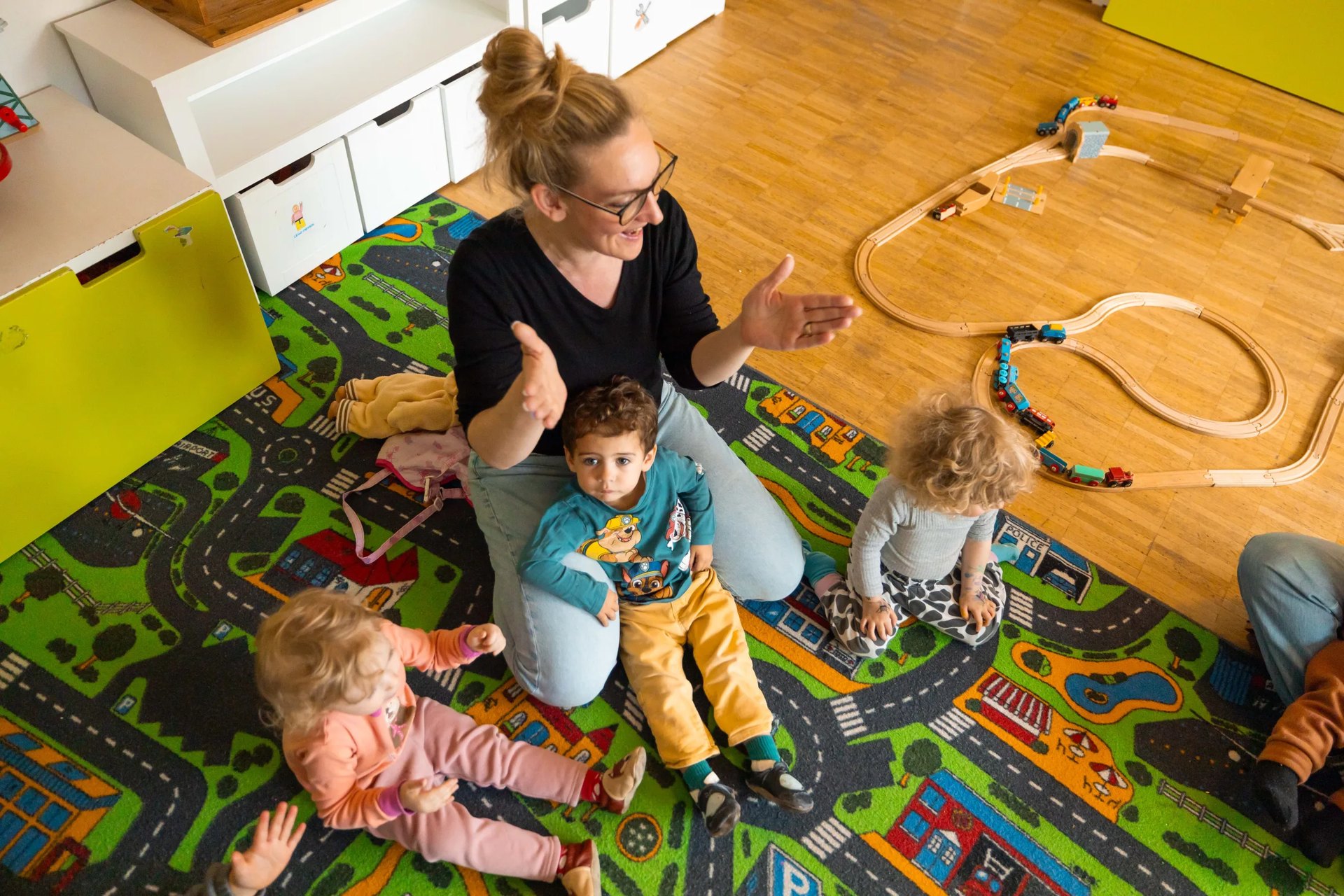 Erzieherin klatscht mit drei Kleinkindern auf dem Spielteppich, von oben fotografiert
