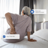 A woman sits on a bed, holding her lower back in pain.