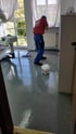 Ein Mann in blauer Hose und rotem Hemd wischt den glänzenden grauen Boden mit einem Mopp; daneben steht ein weißer Kanister.