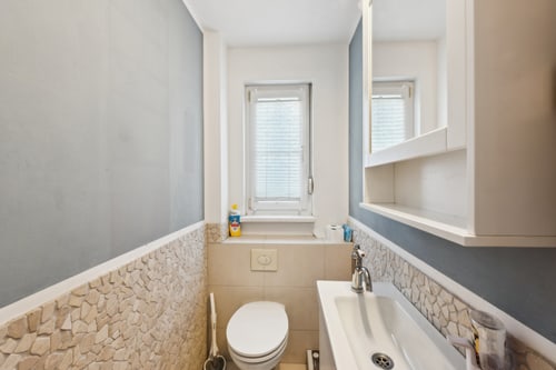 Kleines Gäste-WC mit beigefarbenen Fliesen, weißer Toilette, Waschbecken, Spiegelschrank und Fenster mit Jalousien.