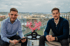 Zwei Männer sitzen auf einem Balkon mit Stadtblick, ein schwarzer Tisch mit rosa Blumen zwischen ihnen.