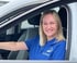 Eine lächelnde Frau mit blonden Haaren und blauem Poloshirt sitzt am Steuer eines weißen Autos.