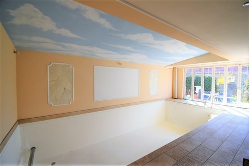 Leeres Hallenbad mit Himmelgemälde an der Decke und großen Fenstern zu einem Garten.