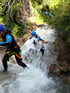 Personen in Neoprenanzügen und Helmen klettern einen felsigen Wasserfall hinunter.