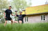 Zwei Männer trainieren im Gras vor einem Haus mit grünem Dach; einer hebt Hanteln, der andere unterstützt.