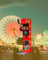 Roter Boxautomat auf einem Jahrmarkt mit Riesenrad und Menschenmenge.