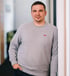 Mann in grauem Pullover mit Levi's Logo und Jeans, mit der Hand in der Hosentasche, lehnt an einer weißen Wand.