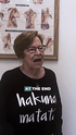An older woman in glasses and a black "Hakuna Matata" shirt speaks, with anatomical charts behind her.